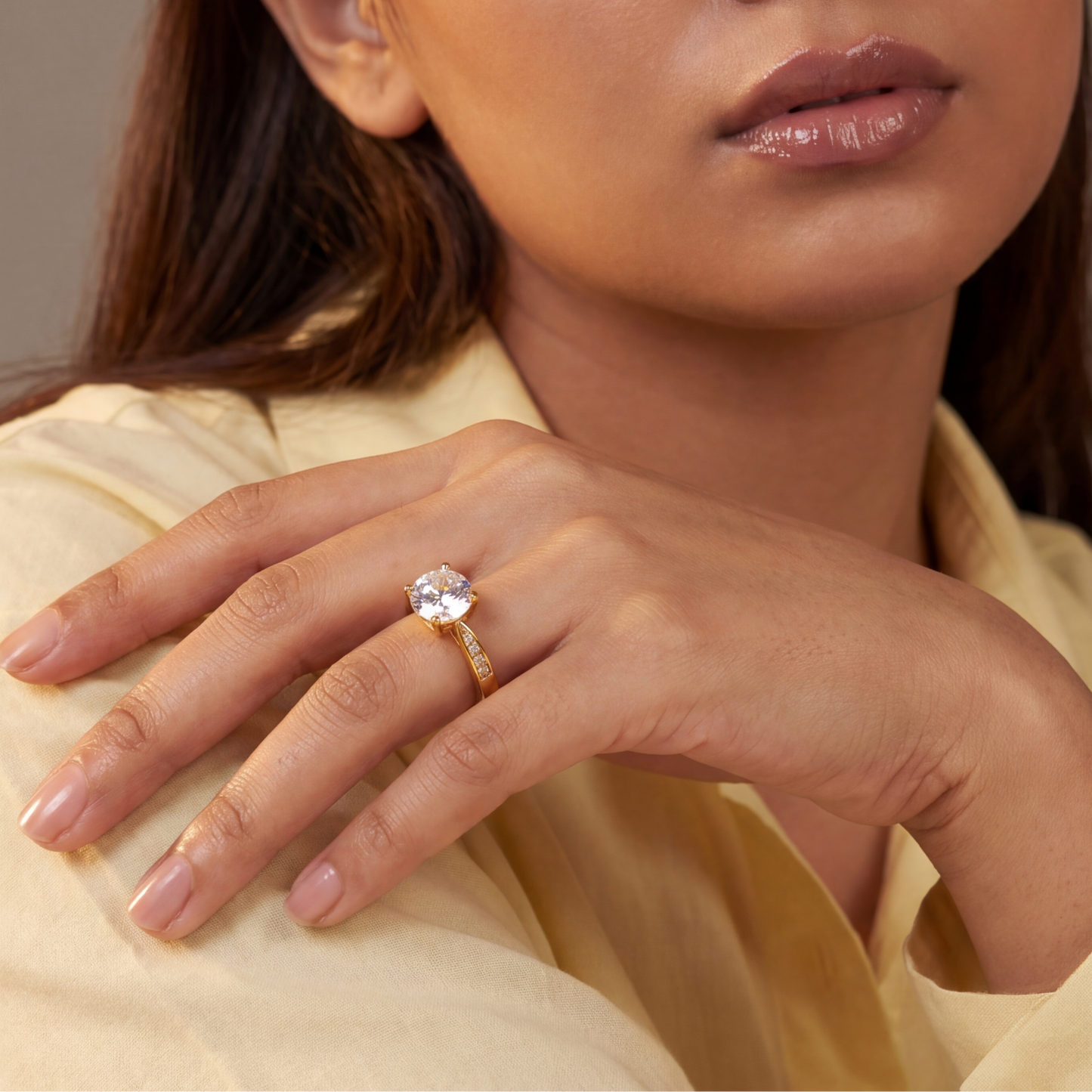 Woman wearing a gold ring with a diamond, posing with one hand on her shoulder against a neutral background.