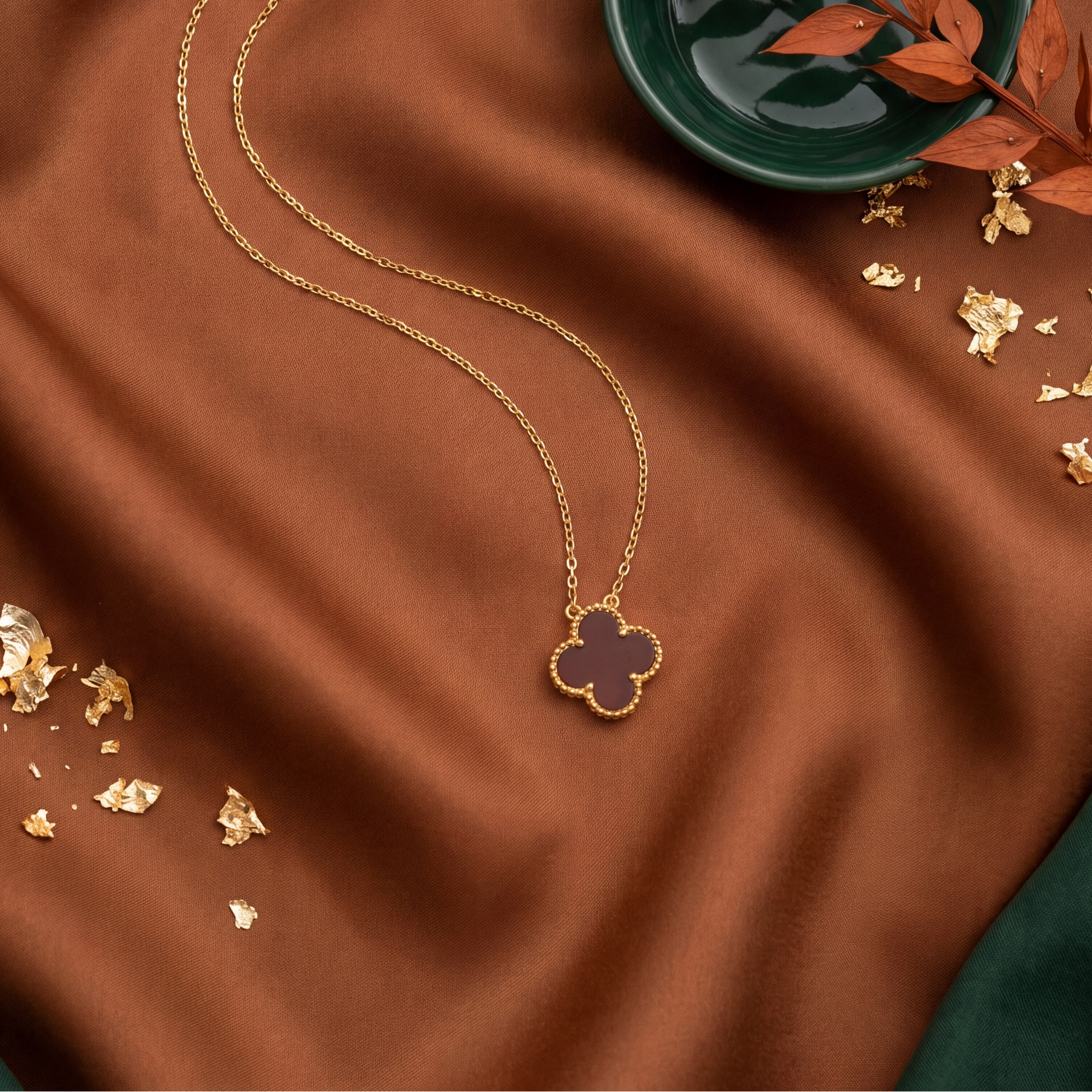 Gold necklace with clover pendant on brown fabric with gold leaves and a green bowl.