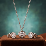 Silver jewelry set with necklace, earrings, and ring on a brown surface with a blurred green background
